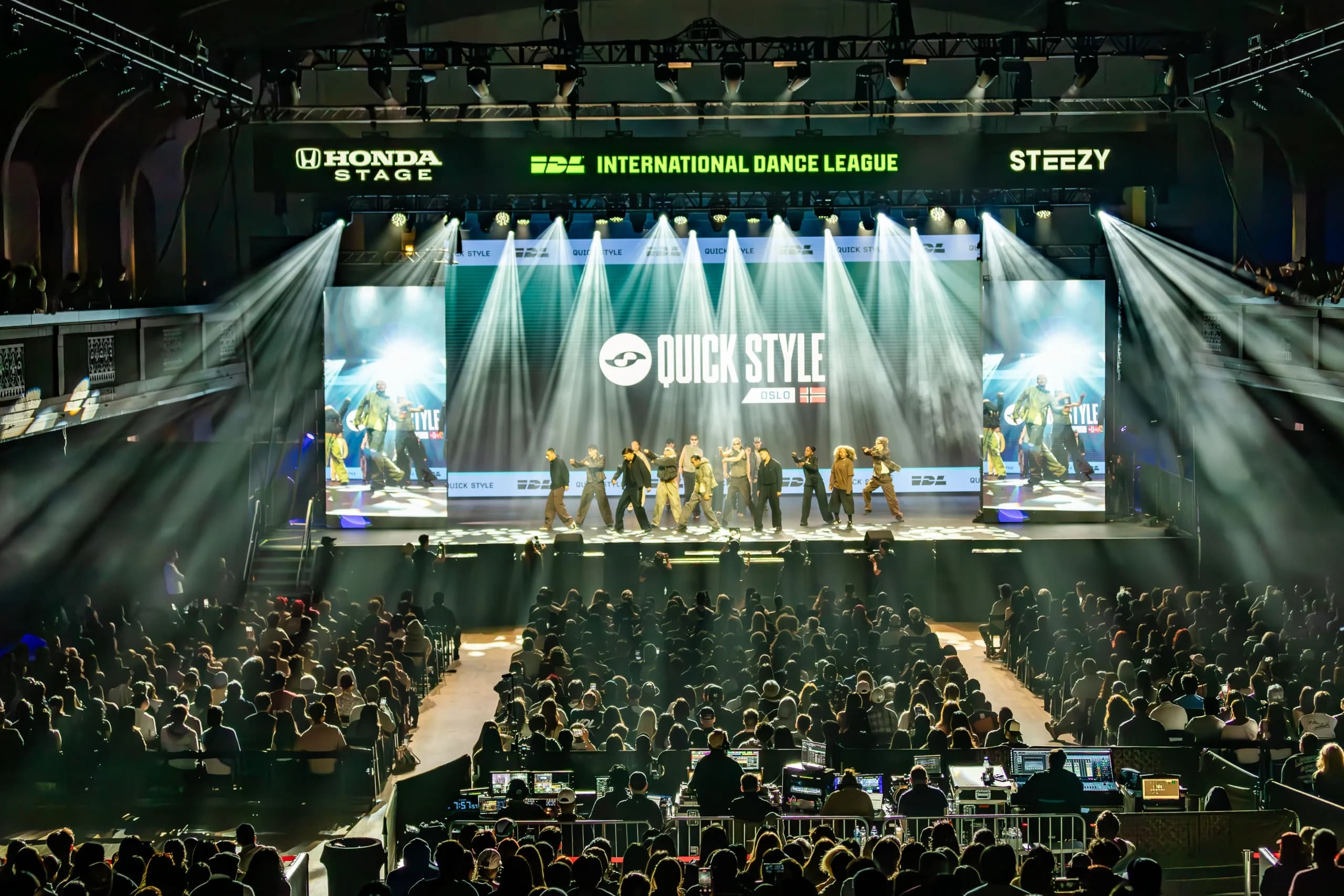 International Dance League main stage — a dance crew performing between two IMAG side screens, the main LED wall carrying the performing team’s name, with a full house and front-of-house consoles in the foreground.