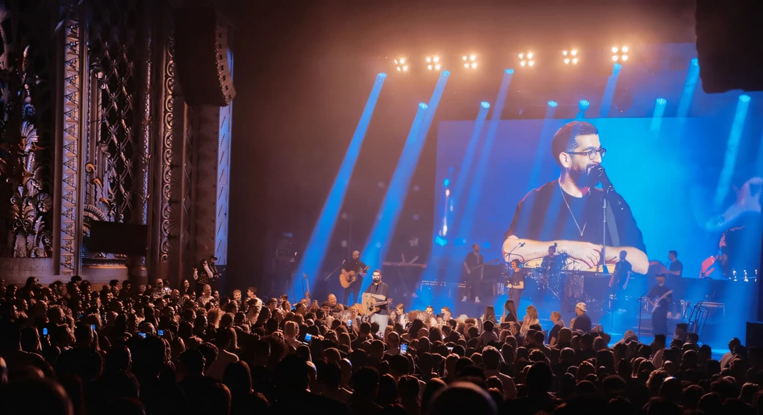 Concert in an ornate historic theatre — a band on stage against a blue LED wall carrying an IMAG close-up of the singer, warm overhead lighting bars and a packed floor.