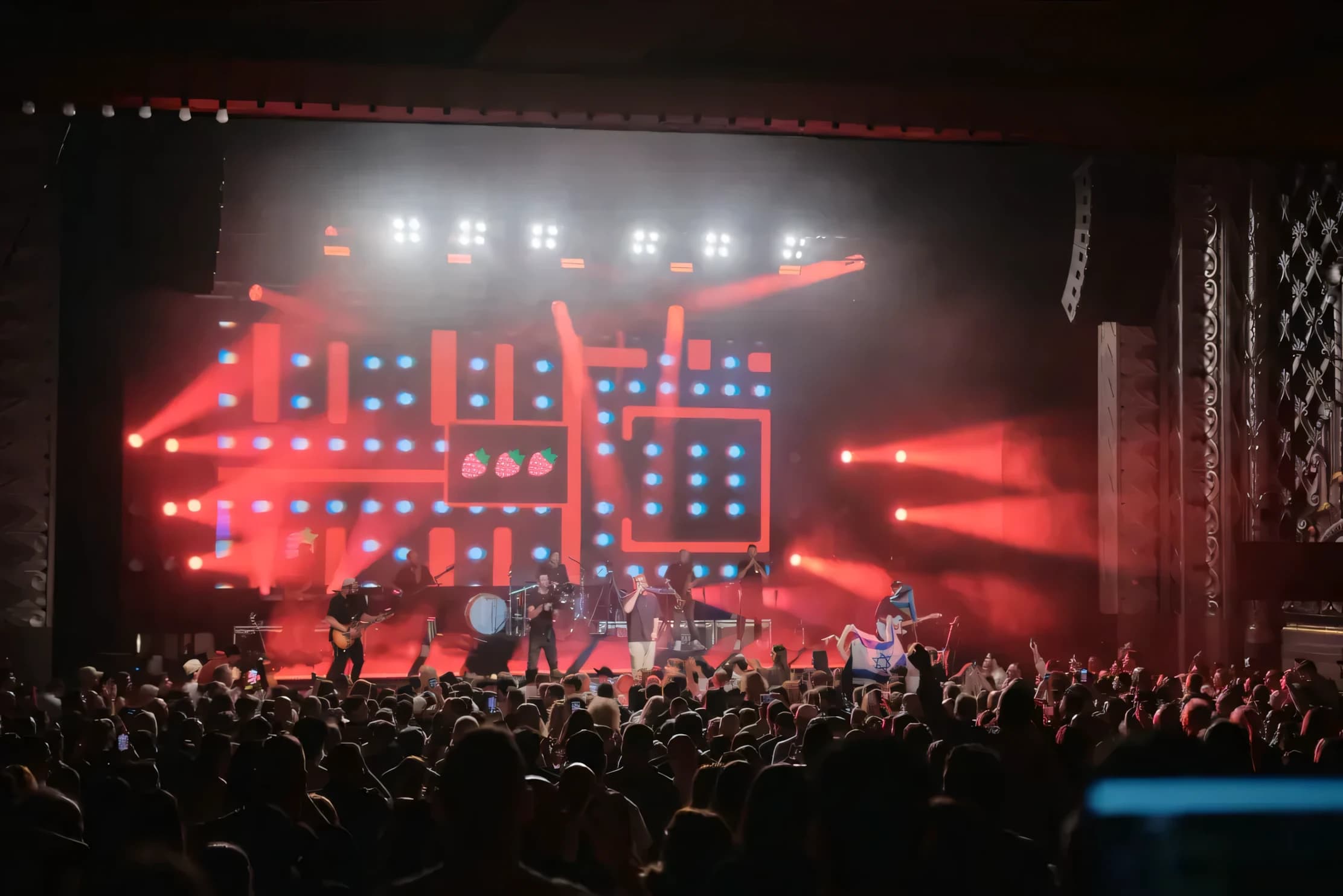 Concert in an ornate theatre under red wash — a band on stage against a large LED wall running graphic content, line array flown stage left, crowd filling the floor.